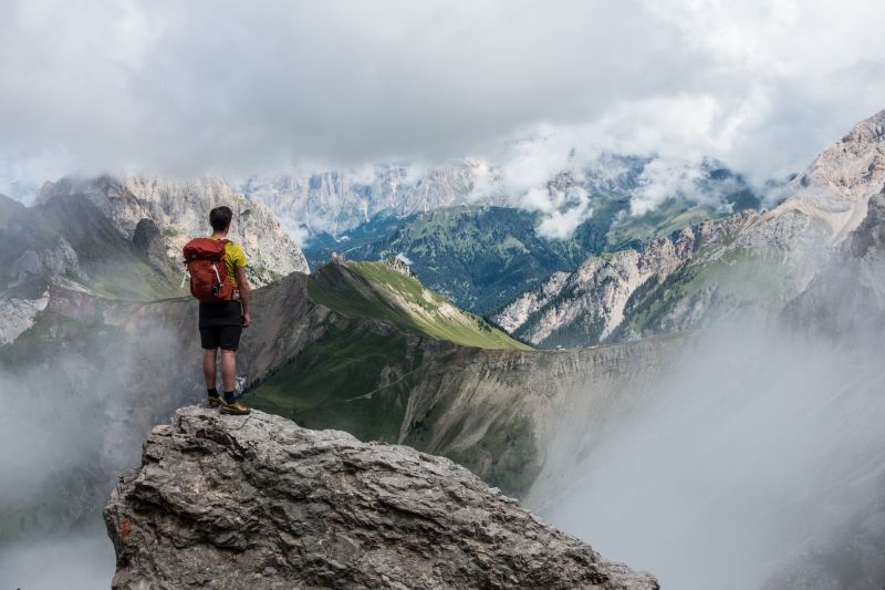 A man with a red backpack stands on a mountaintop looking over more mountains.